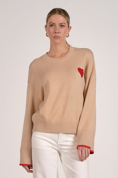 A woman modeling a tan crewneck sweater with a small red heart on the left chest and red trim at the cuffs, paired with white pants, against a light gray studio background.