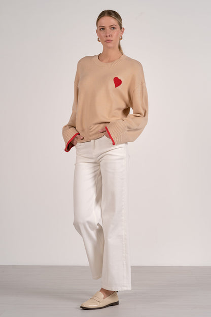 A woman modeling a tan crewneck sweater with a small red heart on the chest and red trim at the cuffs, paired with white wide-leg pants and beige loafers, against a light gray studio background.