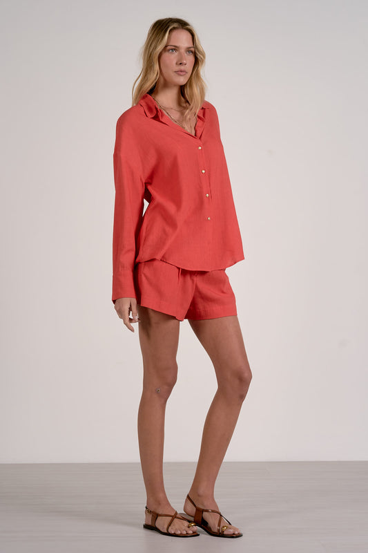 Woman wearing a red outfit with a red button up collared shirt and red shorts with sandals on a plain background