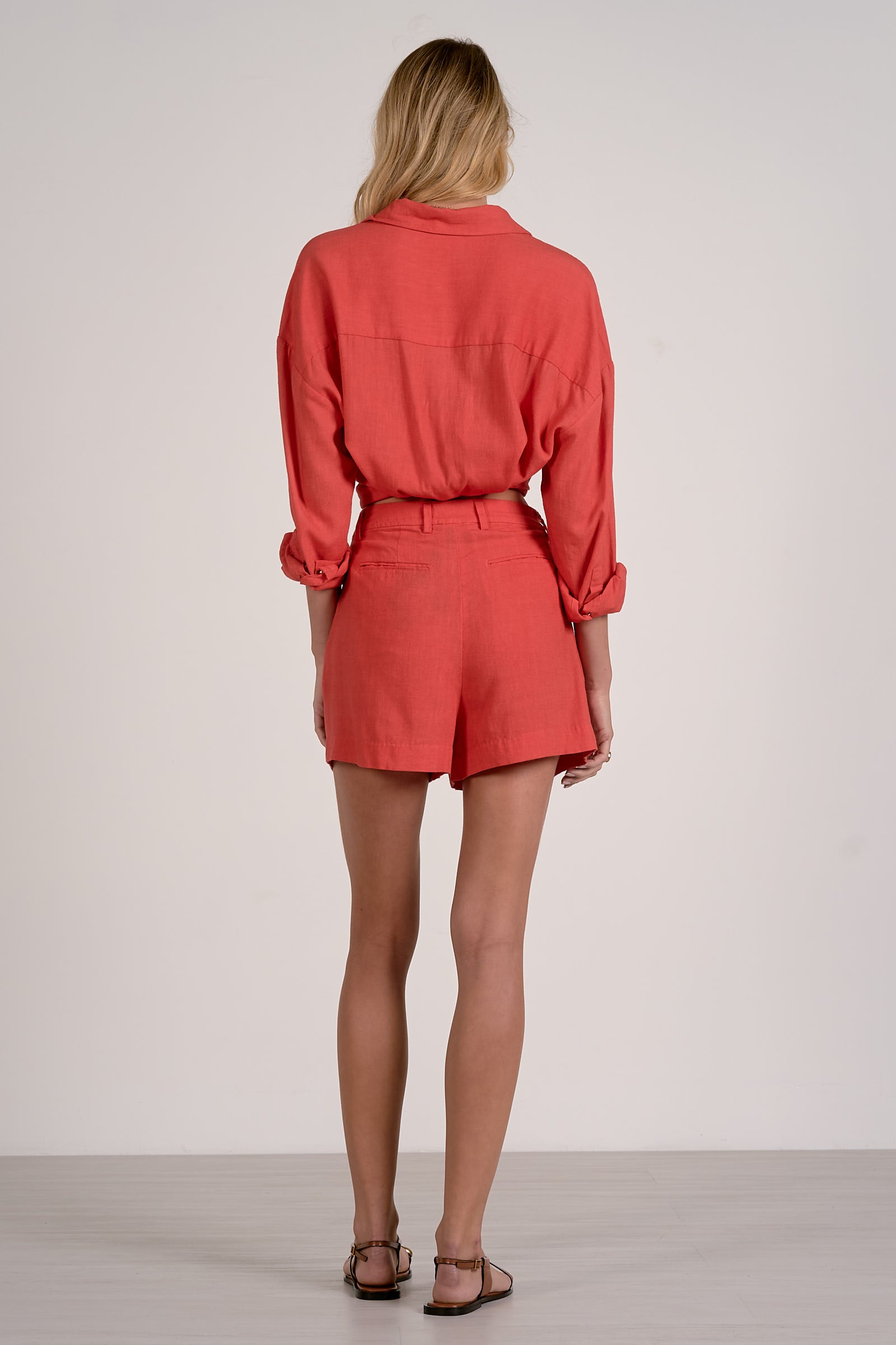 Back view of woman wearing a red top and matching red pocket shorts with sandals on a plain background