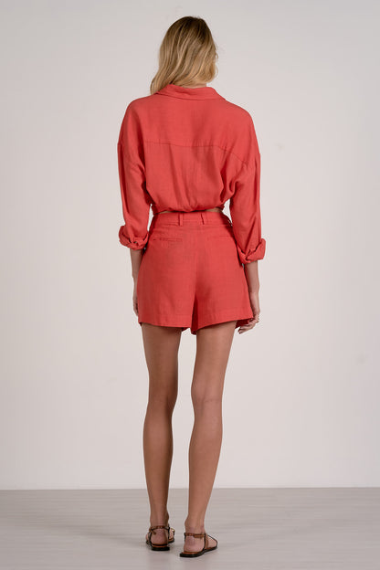 Back view of woman wearing a red top and matching red pocket shorts with sandals on a plain background