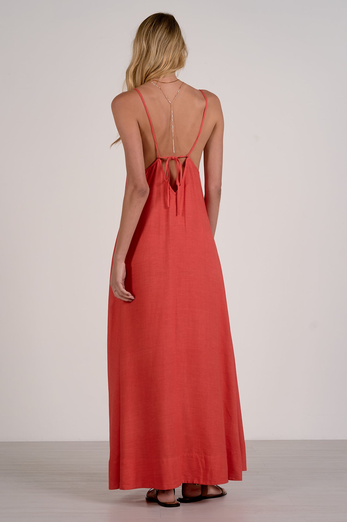 Back view of a woman modeling a red maxi dress from the back, featuring a low open back with a bow detail, set against a plain light gray studio background.