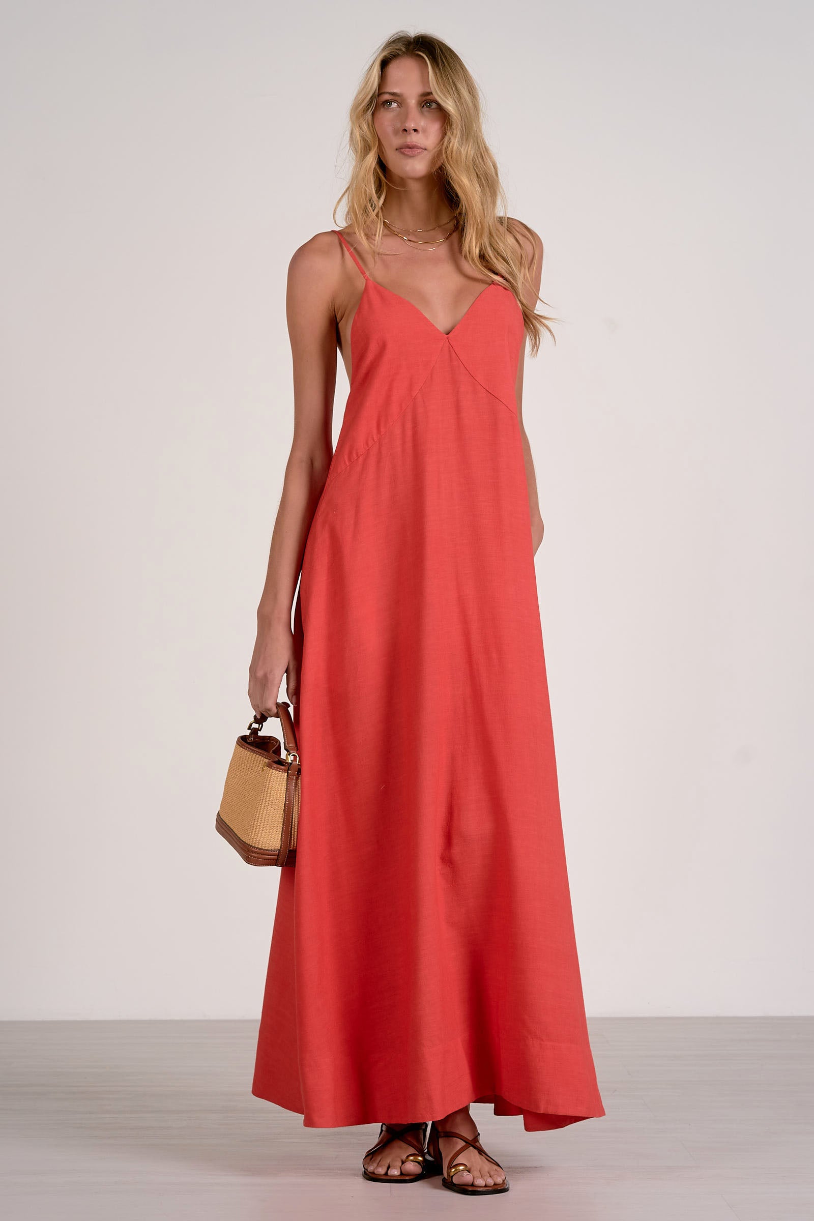 Front view of woman wearing a red v neck maxi dress holding a brown bag on a white background