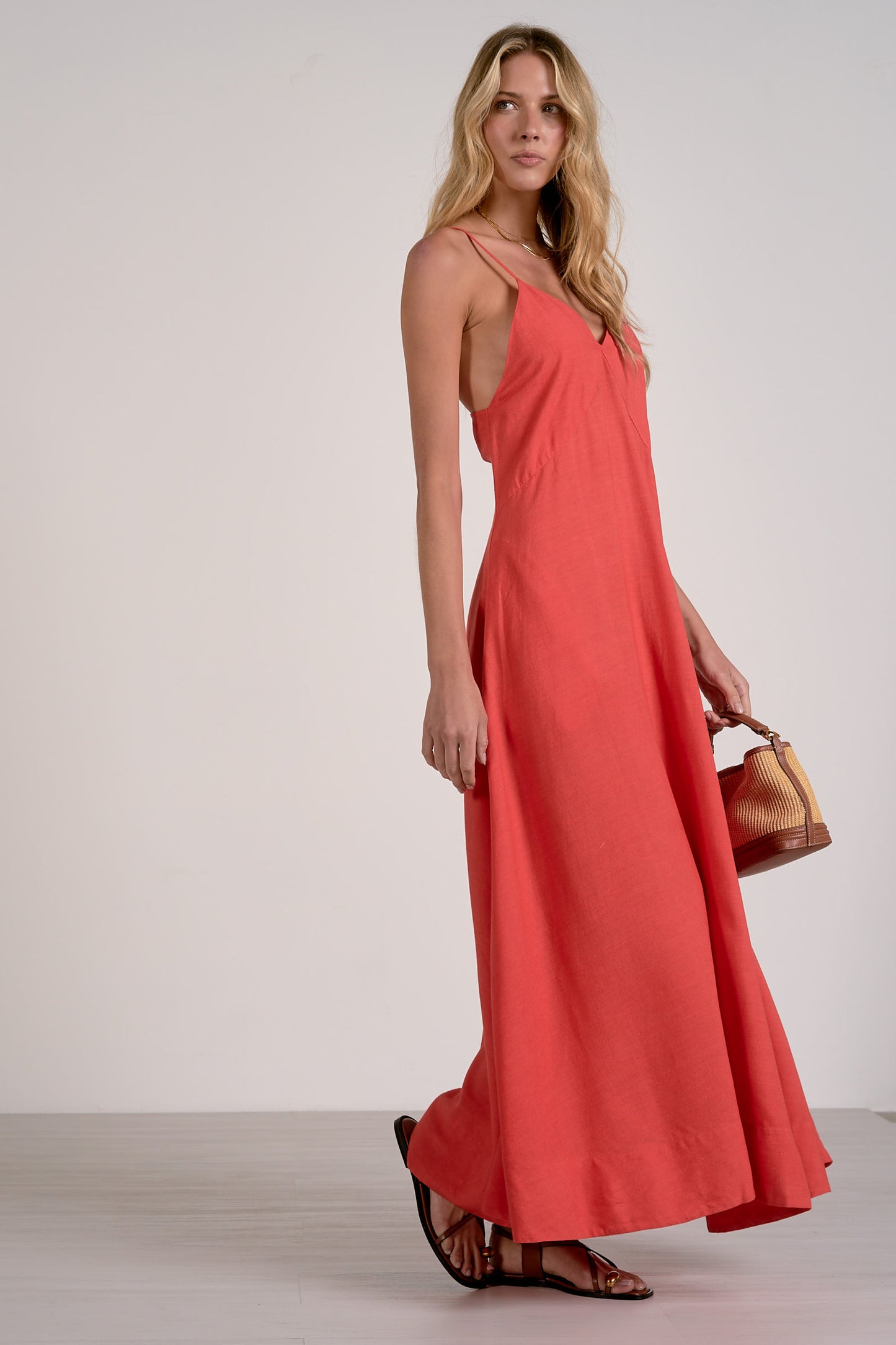Side view of a woman modeling a coral-red maxi dress with thin spaghetti straps, a V-neckline, and a flowing skirt, paired with brown strappy sandals and holding a small brown leather handbag against a plain light gray studio background.
