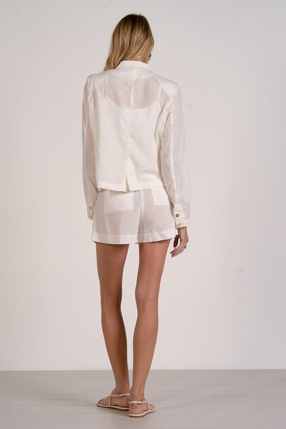 Back view of a woman wearing a white blazer and white shorts with sandals on a plain background. 