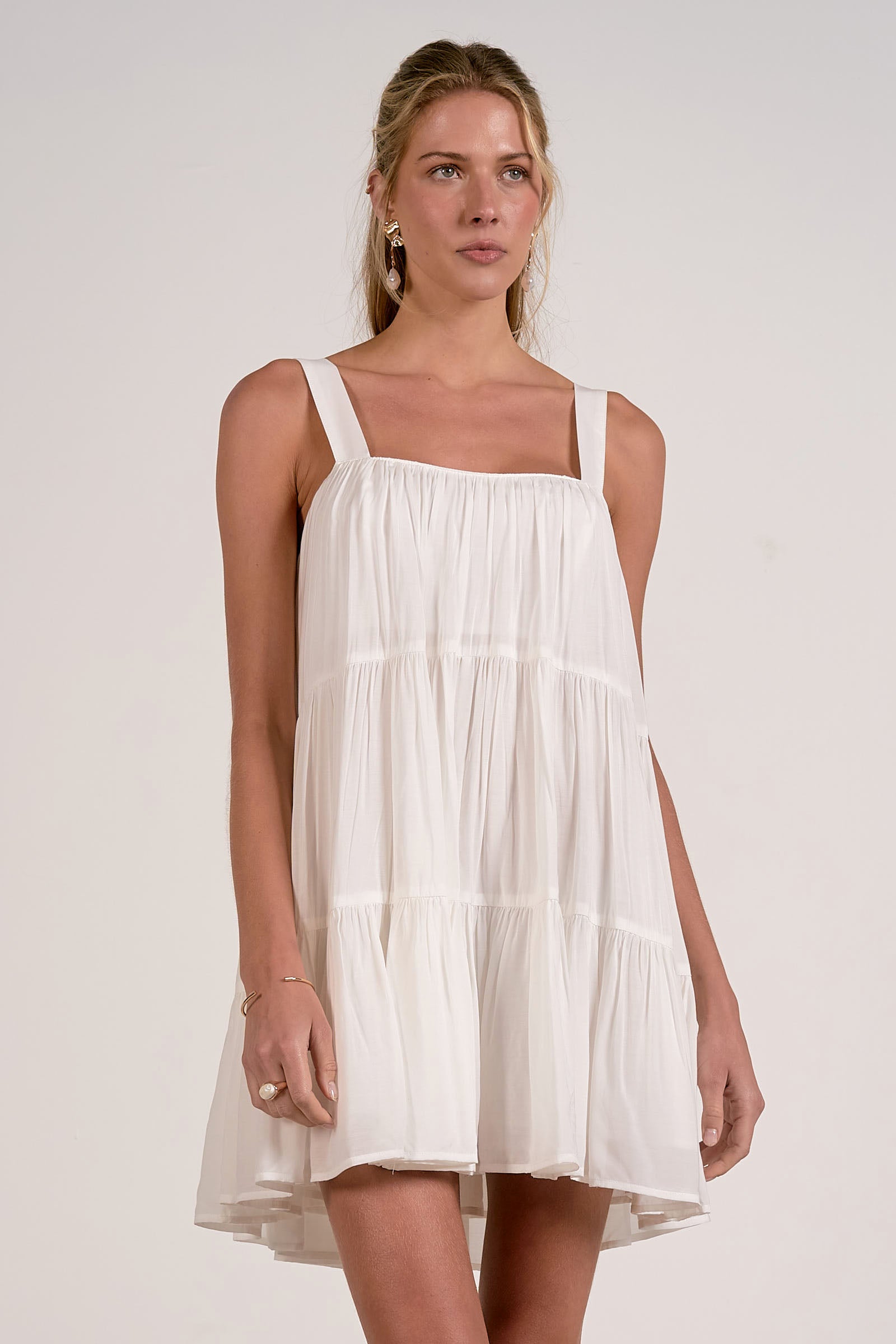 Front view of woman wearing a white tiered mini dress with tank straps against a plain background