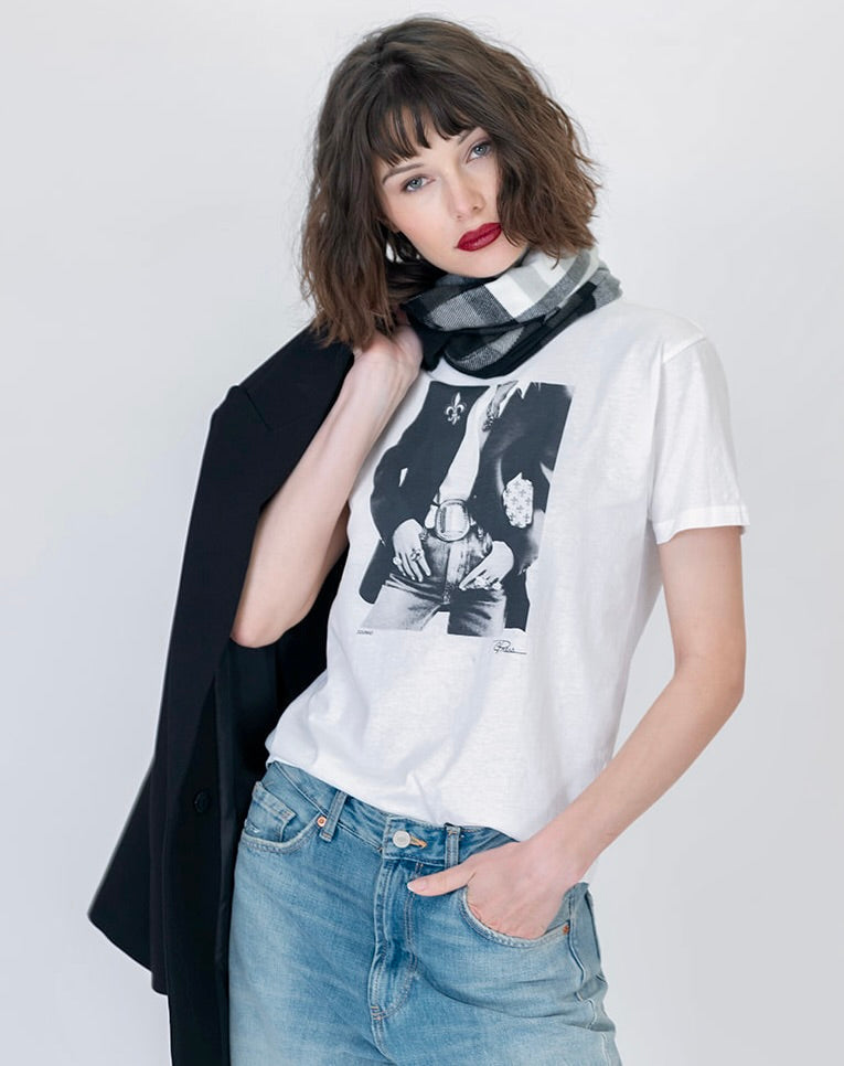 A woman modeling a white graphic t-shirt with a black-and-white iconic print, styled with a black blazer draped over her shoulder, a black and white plaid scarf, and light blue jeans against a light gray studio background.