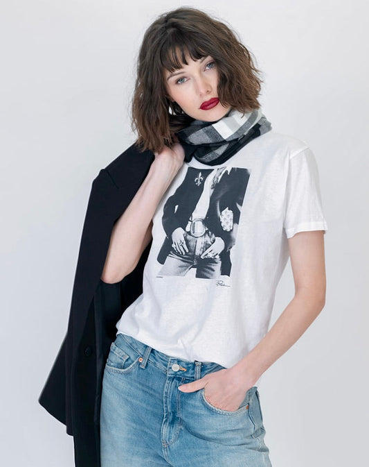 A woman modeling a white graphic t-shirt with a black-and-white iconic print, styled with a black blazer draped over her shoulder, a black and white plaid scarf, and light blue jeans against a light gray studio background.
