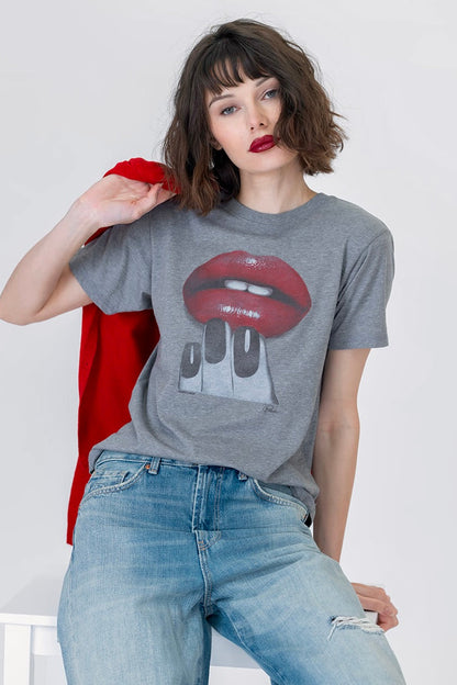 A woman wearing a heather gray graphic t-shirt with a red lips and white teeth and manicured nails design, paired with light blue jeans, holding a red jacket over her shoulder against a white studio background.