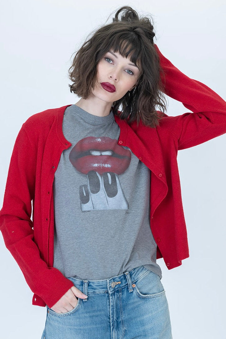 A woman modeling a red button-up cardigan over a gray graphic t-shirt with a red lips & manicure design, paired with blue jeans, against a light gray studio background.