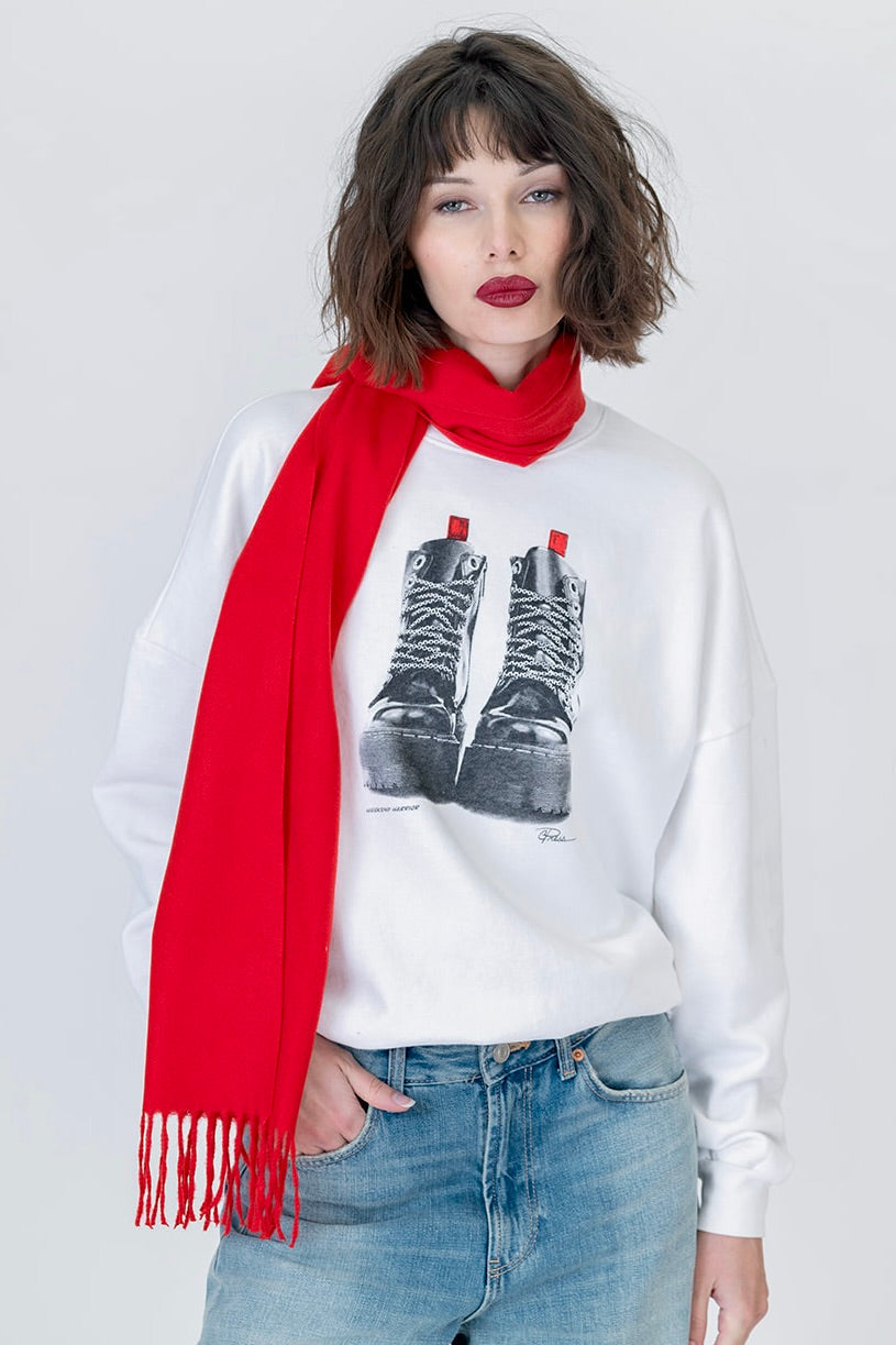 A model wearing a white crewneck "weekend warrior" sweatshirt with a graphic of black lace-up boots, styled with a bright red scarf and blue jeans against a light gray background.