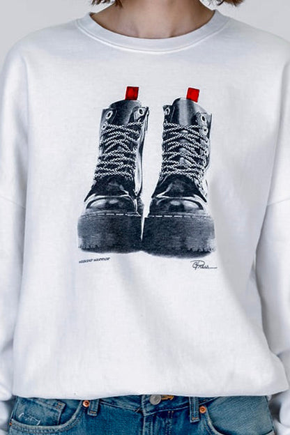 A white crewneck "weekend warrior" sweatshirt featuring a graphic print of a pair of black lace-up combat boots with red pull tabs, worn by a model with blue jeans.