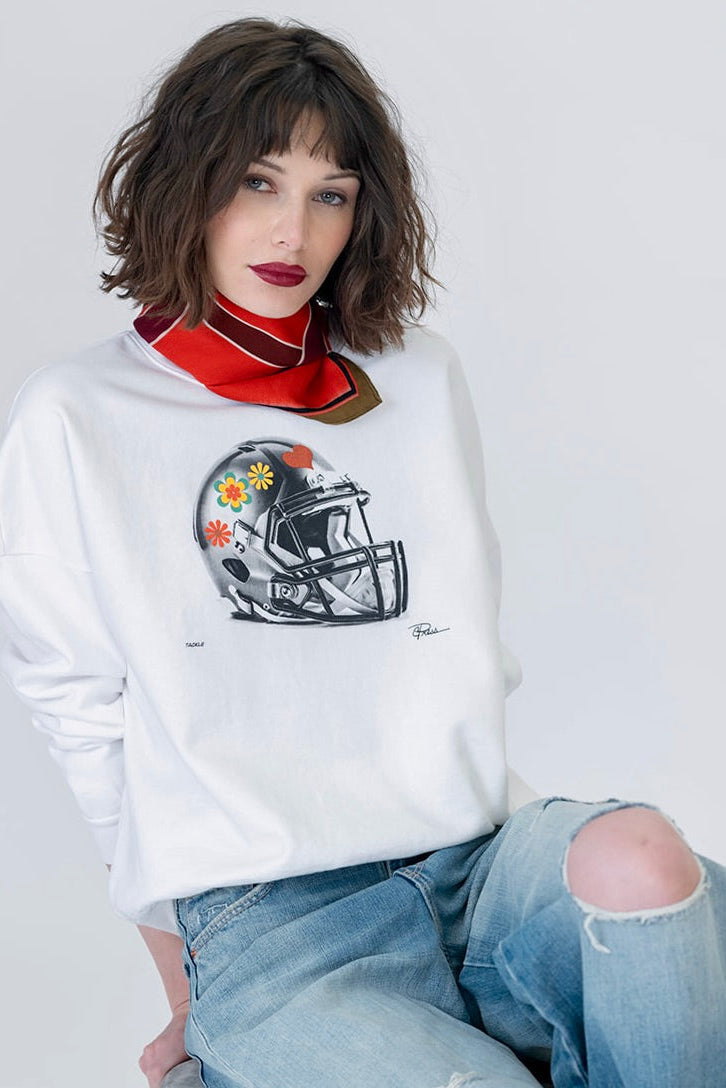 A woman wearing a white crewneck sweatshirt with a graphic of a football helmet on the front, styled with a red and black neck scarf and distressed blue jeans.
