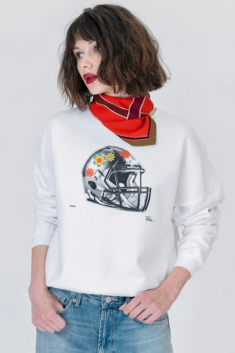 Woman wearing the tackle cozy sweatshirt in white, featuring a football helmet with flowers on it, paired with a red scarf and blue jeans. 