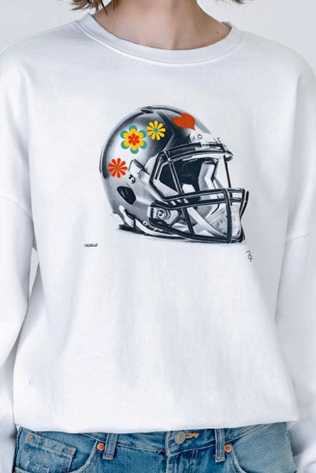 A person wearing a white crewneck sweatshirt with a graphic of a black and gray football helmet decorated with colorful flowers, paired with blue jeans.