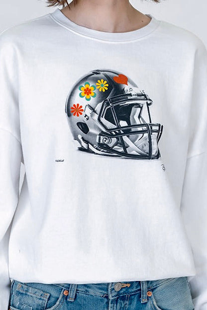 A person wearing a white crewneck sweatshirt with a graphic of a black and gray football helmet decorated with colorful flowers, paired with blue jeans.