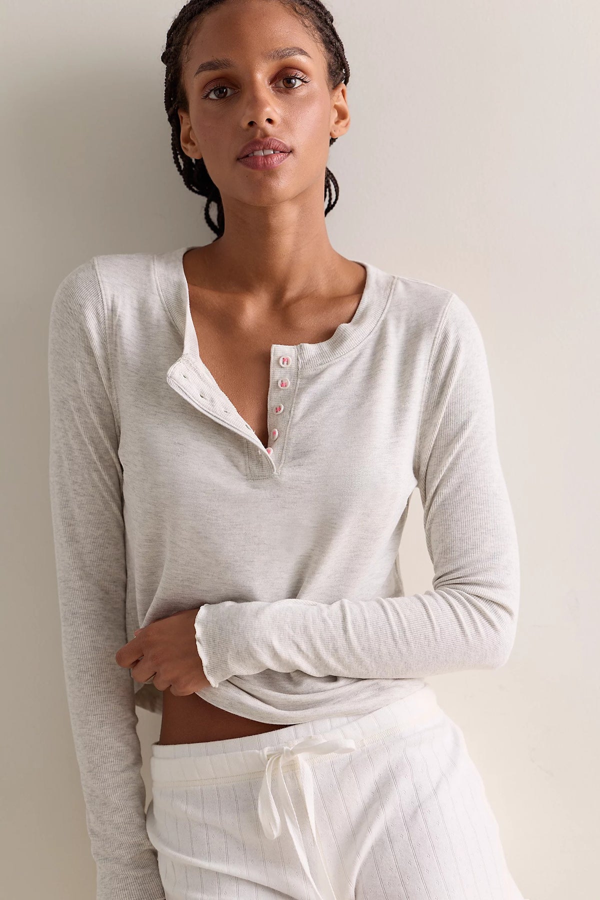 A woman wearing a heather grey long-sleeve henley top with a buttoned split neckline and white drawstring pants, posed against a plain light beige wall.