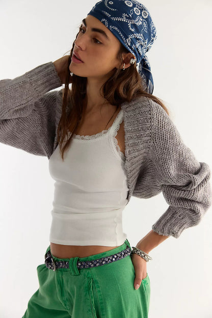 A woman wearing a gray opal shrug sweater, a white ribbed camisole, a blue paisley bandana, and green pants with a patterned belt, photographed against a plain light background.