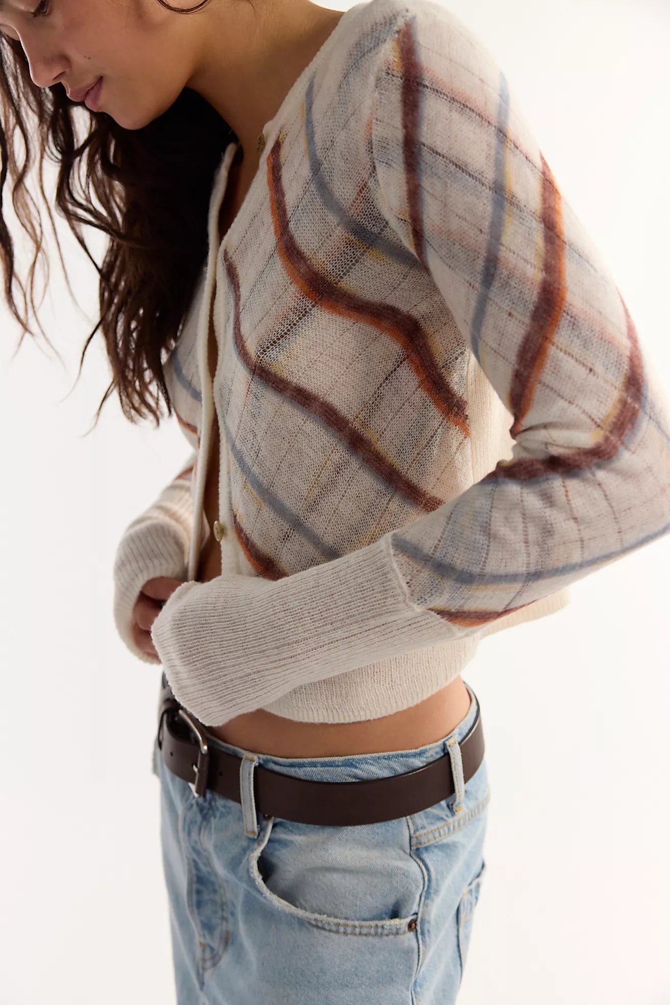 A woman wearing a cropped, long-sleeve knit cardigan with a beige, brown, sugar mist combo plaid pattern, paired with light-wash blue jeans and a dark brown belt, against a plain white background.