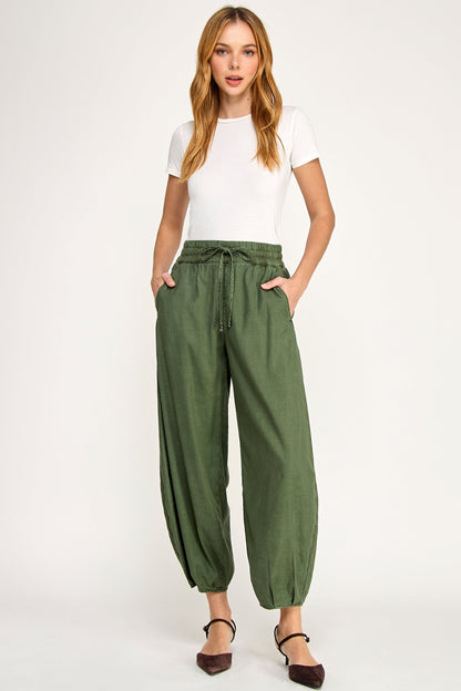 A woman modeling a pair of avocado green wide-leg ellie mini balloon pants with a drawstring waist, paired with a white t-shirt and dark brown flats, against a plain white studio background.