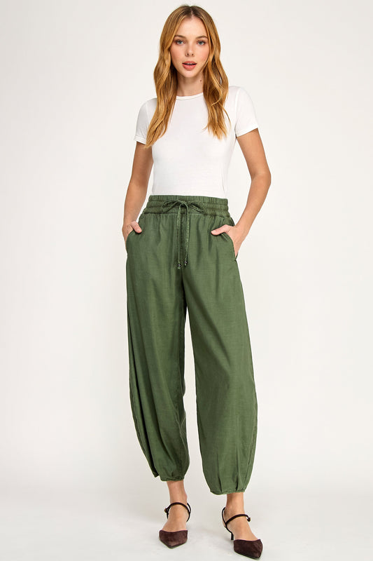 A woman modeling a pair of avocado green wide-leg ellie mini balloon pants with a drawstring waist, paired with a white t-shirt and dark brown flats, against a plain white studio background.