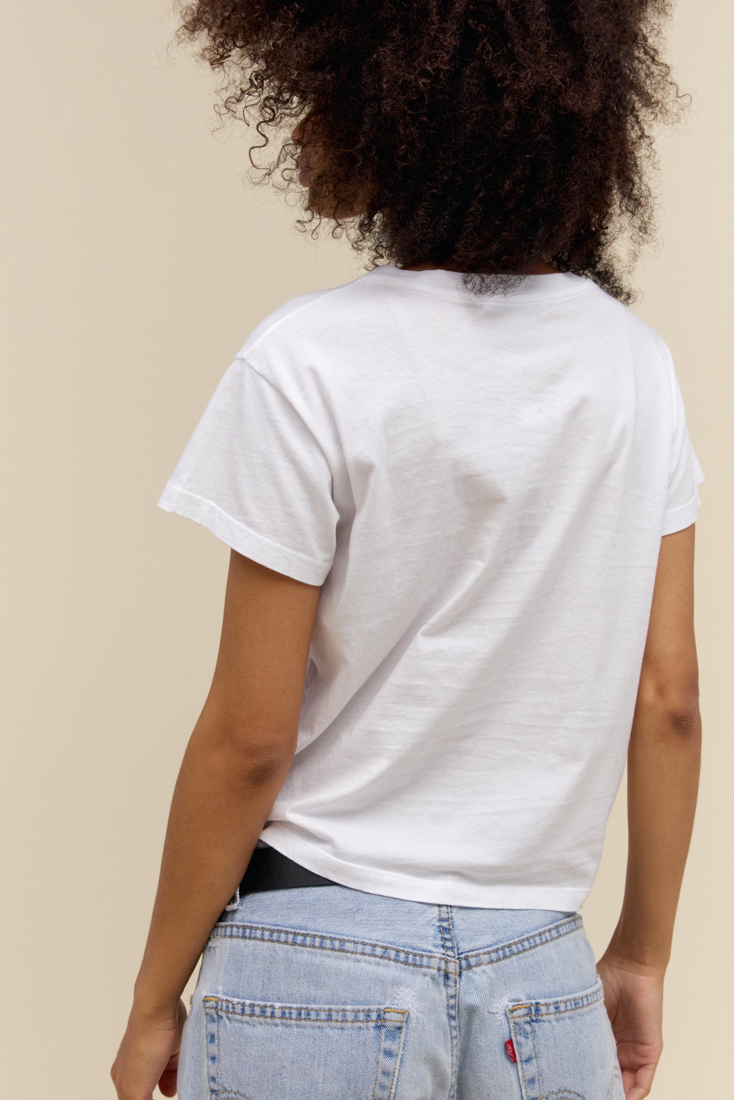 Back view of person wearing a bleach white t-shirt and light blue jeans against a beige background
