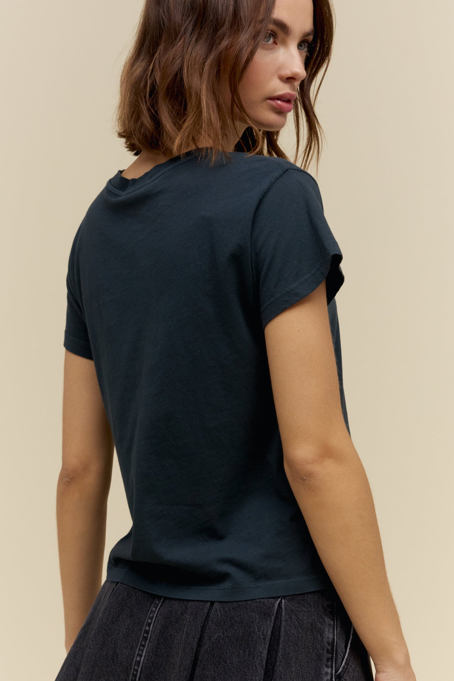 A woman shown from the back wearing a vintage black short-sleeve t-shirt and a dark pleated skirt, set against a plain beige background.