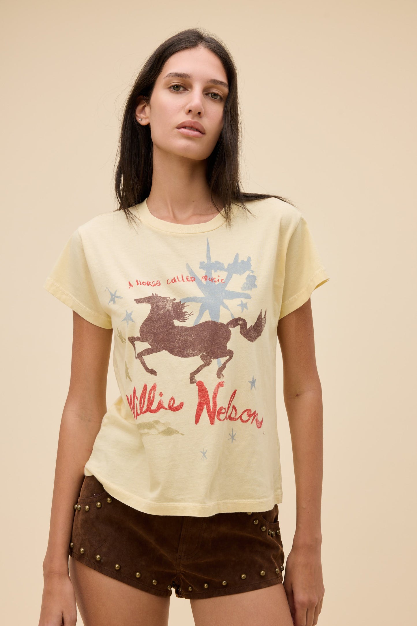 A woman modeling a sheer yellow graphic t-shirt with a brown horse and red 'Willie Nelson' text, paired with brown studded shorts, against a solid beige background.