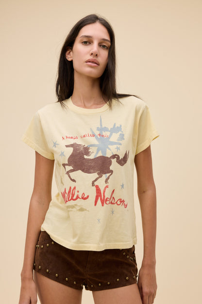 A woman modeling a sheer yellow graphic t-shirt with a brown horse and red 'Willie Nelson' text, paired with brown studded shorts, against a solid beige background.
