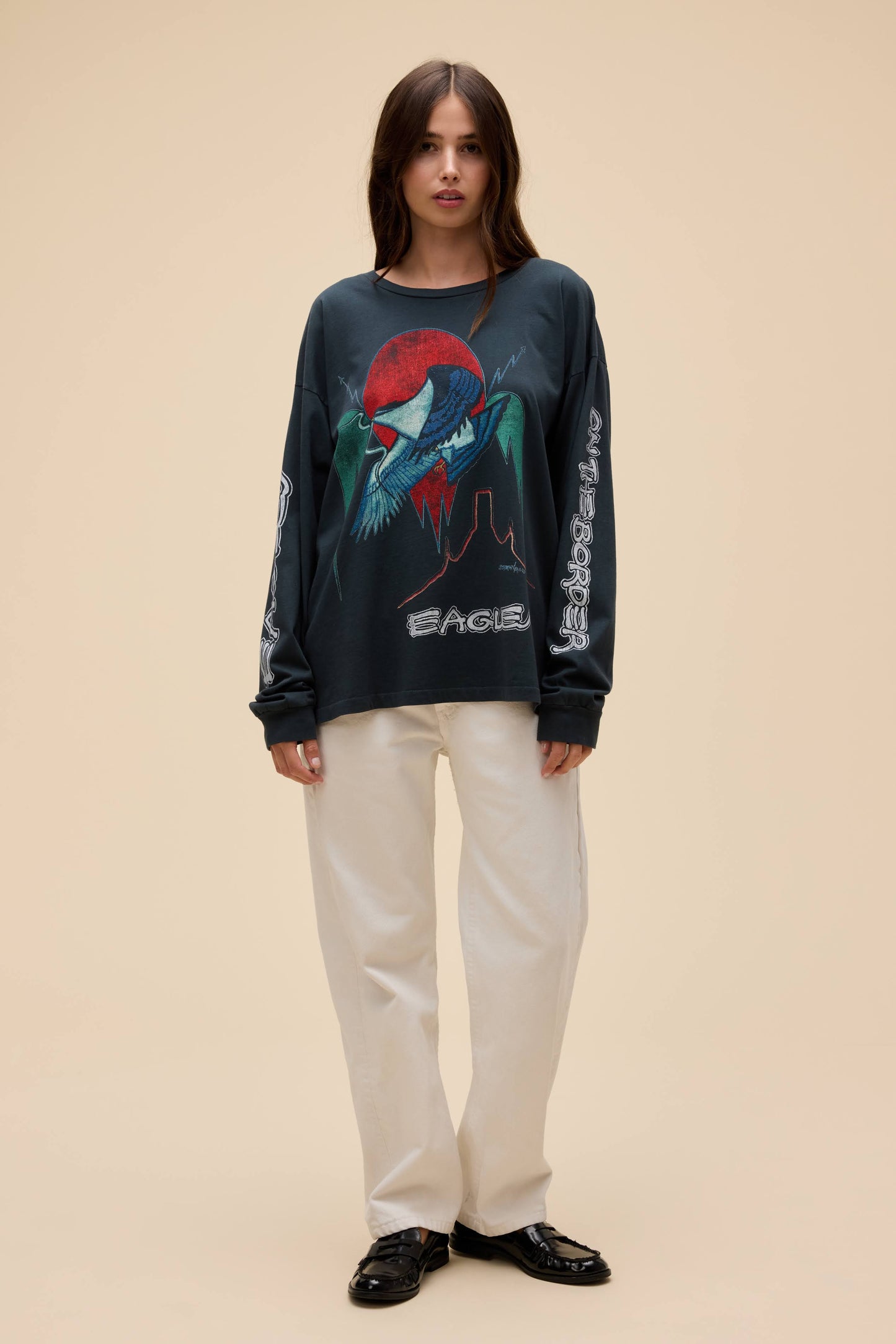 A model stands against a solid beige background wearing a vintage black Eagles graphic long-sleeve shirt with a red, green, and white eagle design, paired with off-white trousers and black loafers.