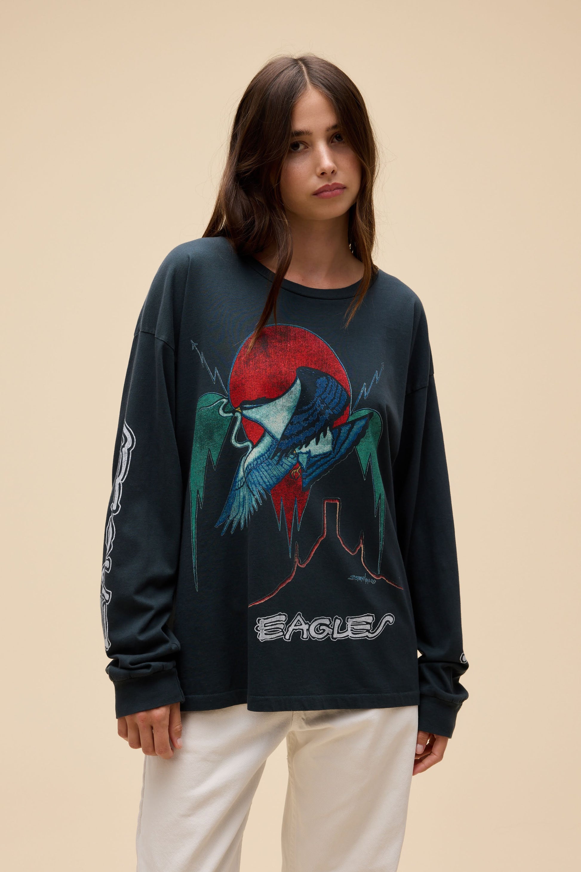 A woman modeling a vintage black long-sleeve graphic sweatshirt featuring a stylized Eagles design in red, blue, and green, paired with off-white pants, against a solid beige background.