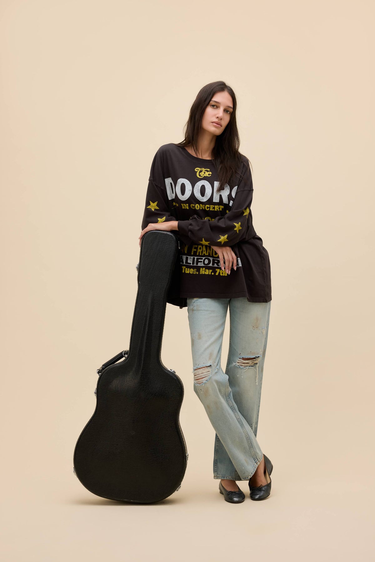A woman stands in a studio wearing a "The Doors" black graphic long-sleeve shirt with yellow and white text, light-wash distressed jeans, and black flats, while leaning against a black guitar case. The background is a solid warm beige.