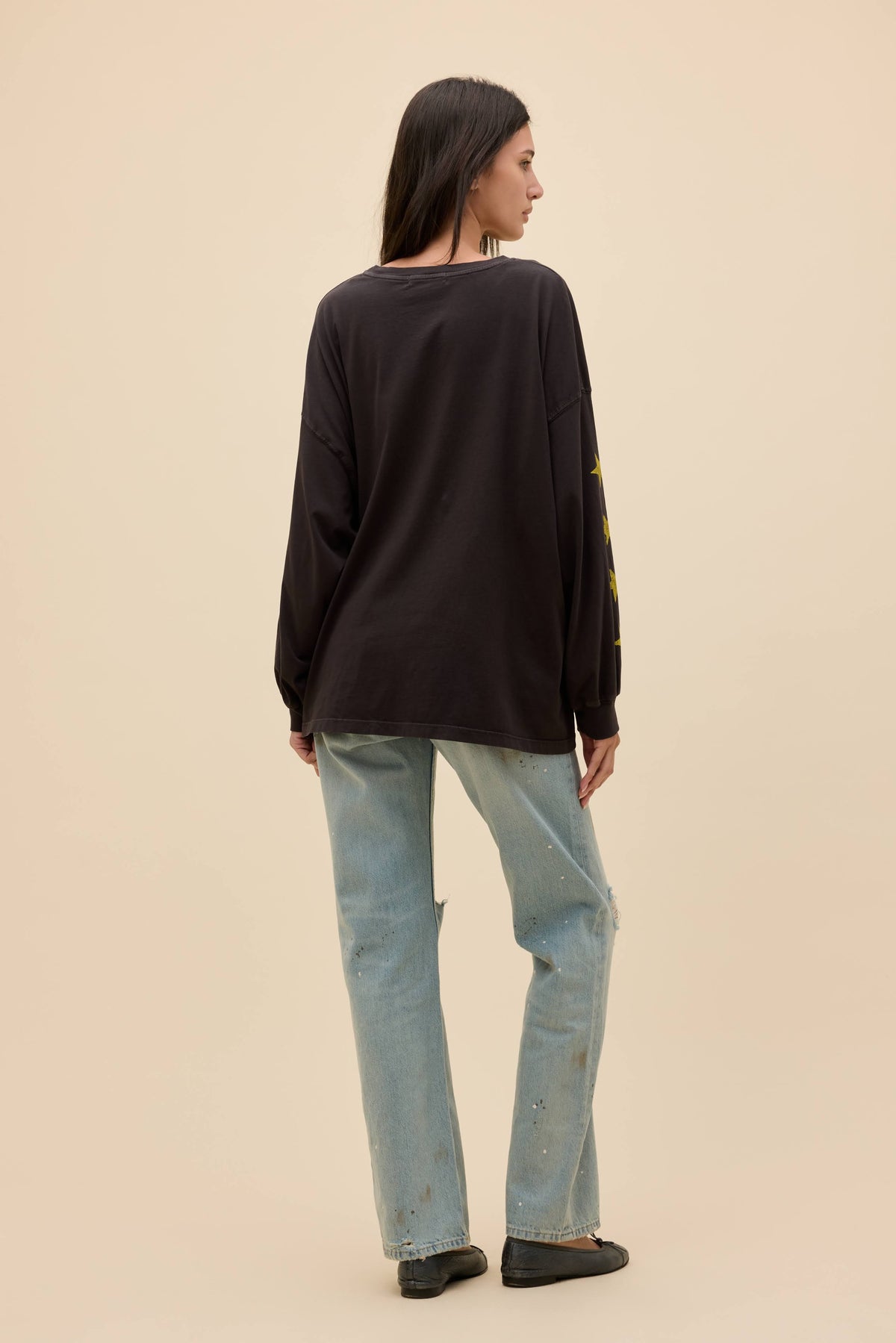 A woman is shown from the back wearing a black long-sleeve top with yellow graphic details on the sleeve, paired with light-wash blue jeans and black flats, set against a solid beige background.