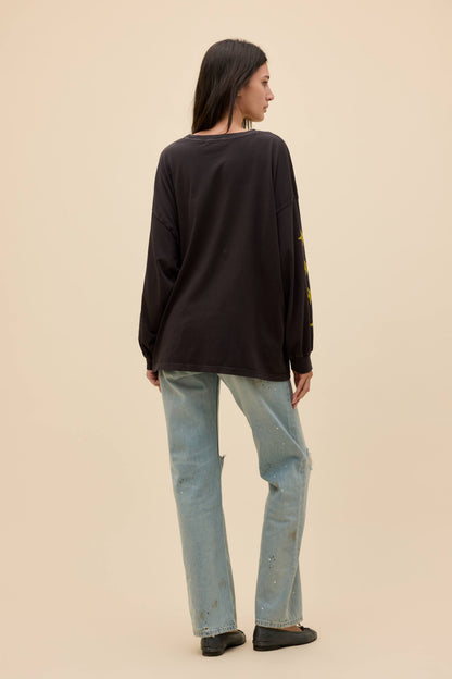 A woman is shown from the back wearing a black long-sleeve top with yellow graphic details on the sleeve, paired with light-wash blue jeans and black flats, set against a solid beige background.