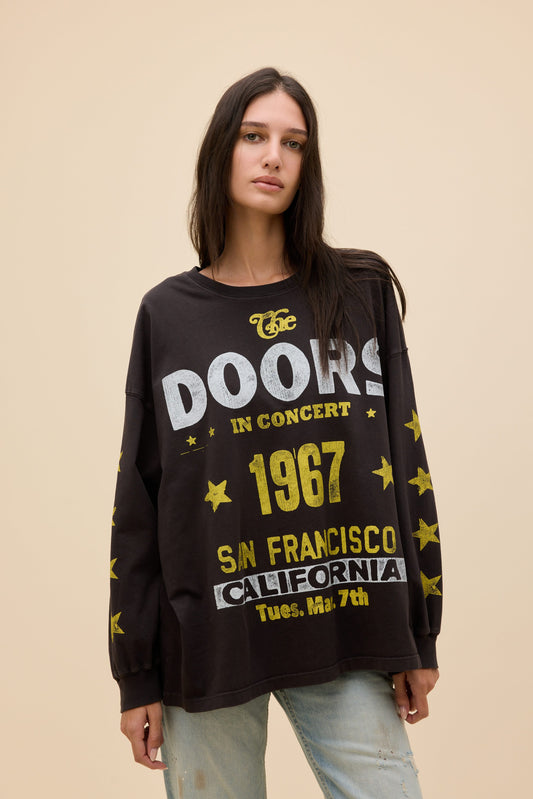 A woman modeling a black long-sleeve graphic sweatshirt with 'The Doors In Concert 1967 San Francisco California' printed in white and yellow on the front, paired with light-wash jeans against a solid beige background.