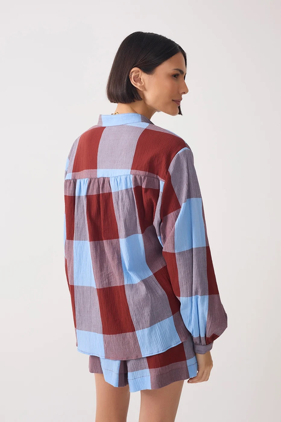 A woman shown from the back wearing a loose, long-sleeve plaid shirt and matching shorts in a dusty red, light blue, and lavender-gray colorway, set against a plain off-white studio background.