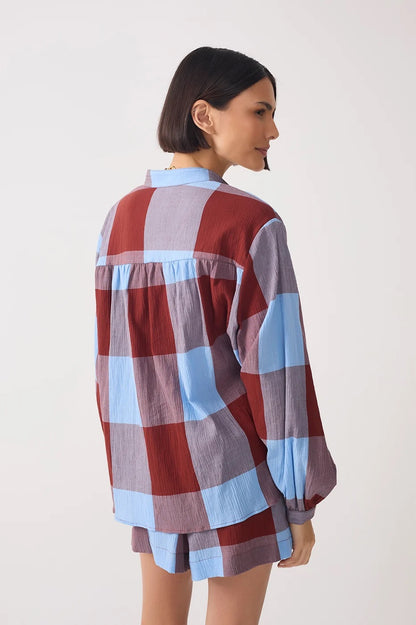 A woman shown from the back wearing a loose, long-sleeve plaid shirt and matching shorts in a dusty red, light blue, and lavender-gray colorway, set against a plain off-white studio background.