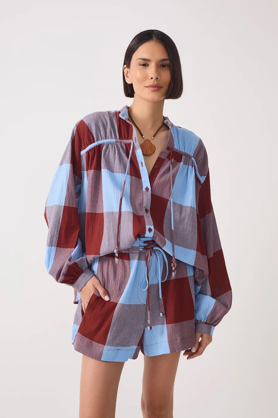 A woman modeling a matching two-piece plaid lounge set with a long-sleeve button-front top and coordinating shorts in dusty red, light blue, and mauve tones, against a plain light gray studio background.