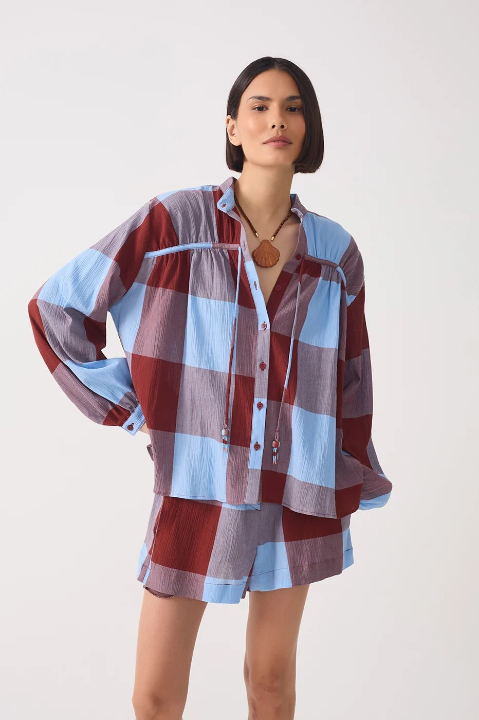 A model wearing a matching plaid lounge set consisting of a long-sleeve button-up shirt and shorts. The fabric features a large checkered pattern of light blue, dusty purple, and deep burgundy red.