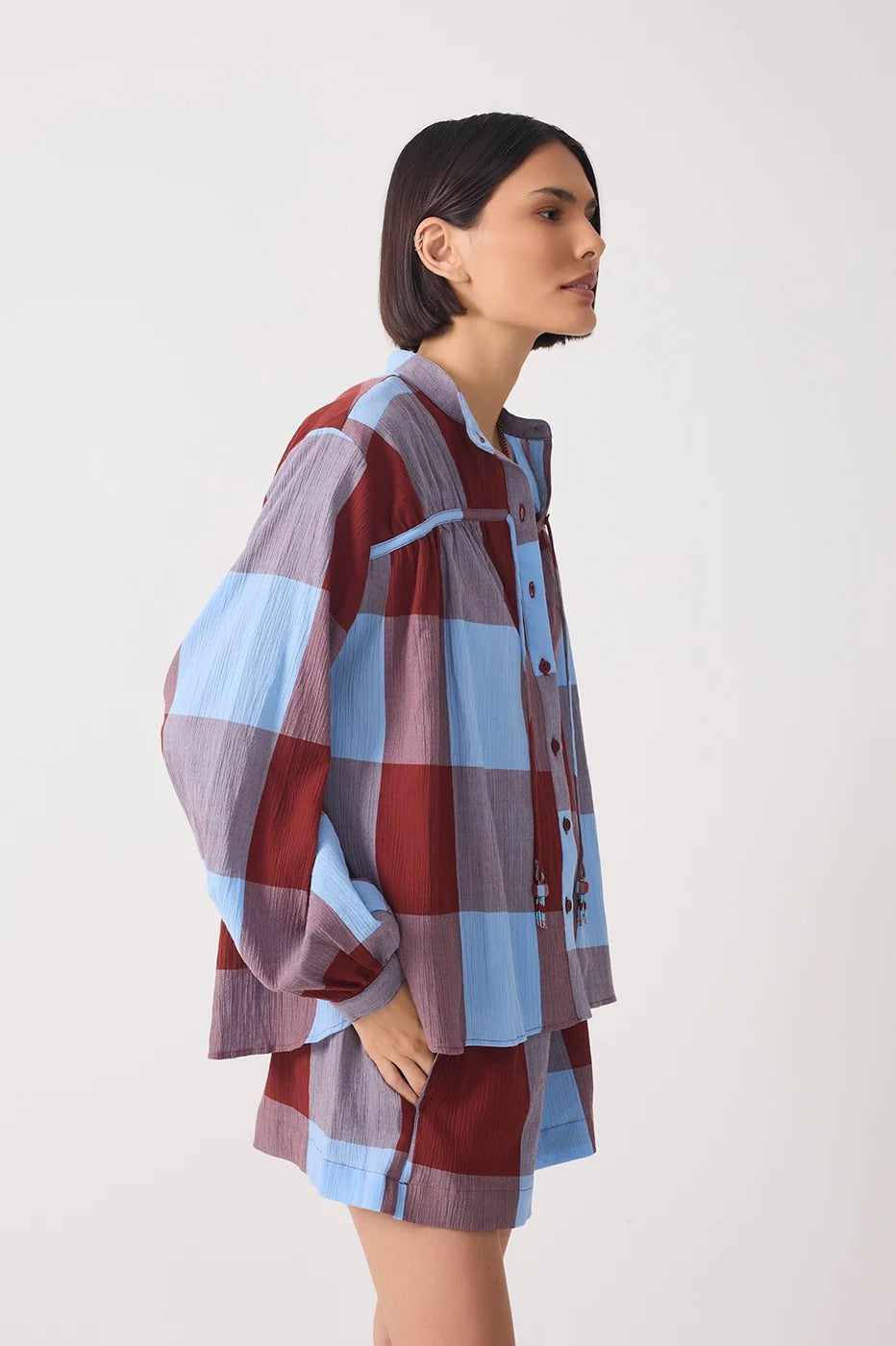 A woman wearing a loose, oversized plaid shirt and matching shorts in dusty red, light blue, and muted purple, photographed against a plain light gray studio background.