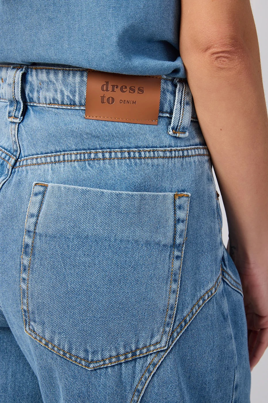 Close-up of the back waistband of light blue denim jeans, showing a brown leather patch with the brand name 'dress to DENIM' and a large back pocket.