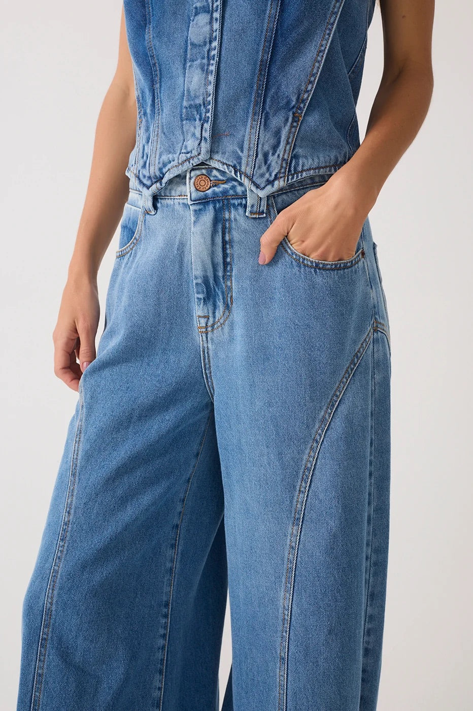 Close-up of a person wearing light blue denim jeans with wide legs, front pockets, and a button closure, against a plain light background.