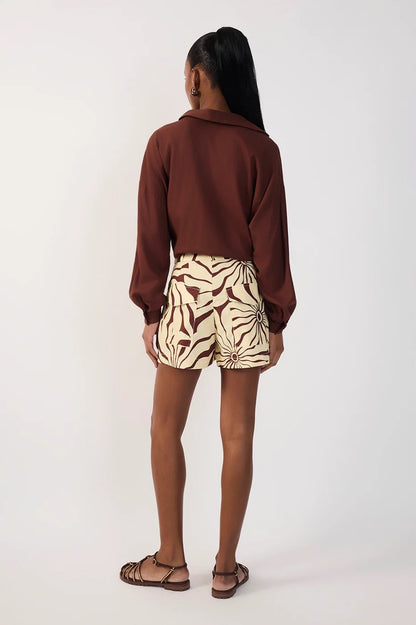 A woman is shown from behind wearing a brown long-sleeve collared shirt and high-waisted shorts with a beige and brown alvorecer pattern, paired with brown strappy sandals, against a plain white studio background.