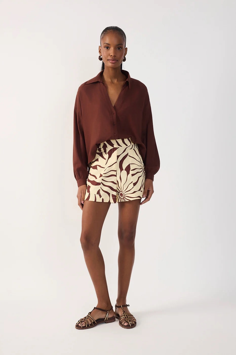 A model stands against a plain light gray studio background wearing a long-sleeve brown collared shirt, a high-waisted beige and brown patterned alvorecer shorts, and brown strappy sandals.