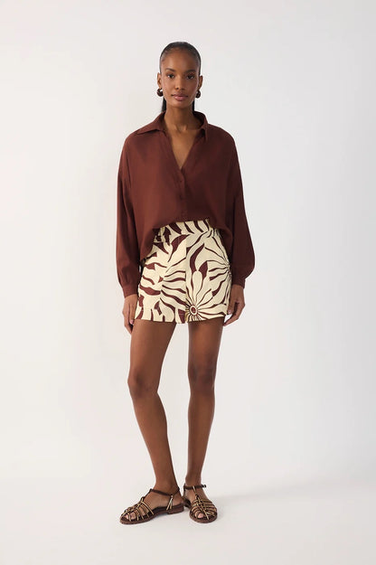 A model stands against a plain light gray studio background wearing a long-sleeve brown collared shirt, a high-waisted beige and brown patterned alvorecer shorts, and brown strappy sandals.