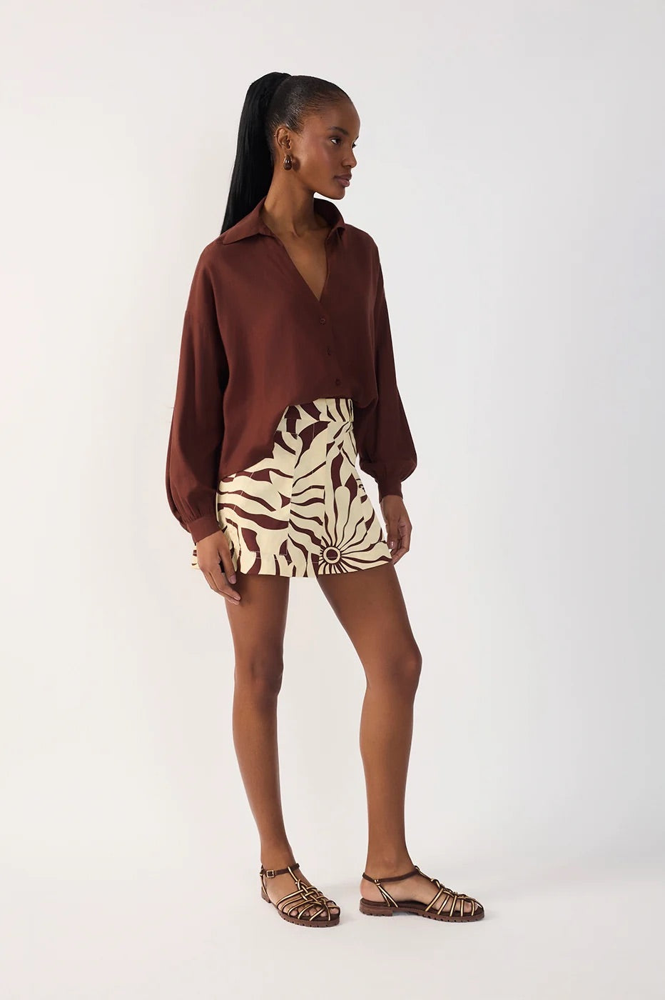 A woman modeling a brown long-sleeve collared shirt and a beige and brown alvorecer patterned shorts paired with brown strappy sandals, against a plain white studio background.