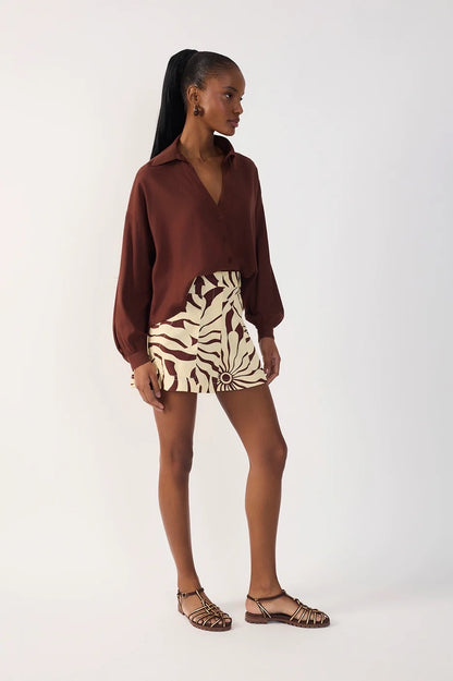 A woman modeling a brown long-sleeve collared shirt and a beige and brown alvorecer patterned shorts paired with brown strappy sandals, against a plain white studio background.
