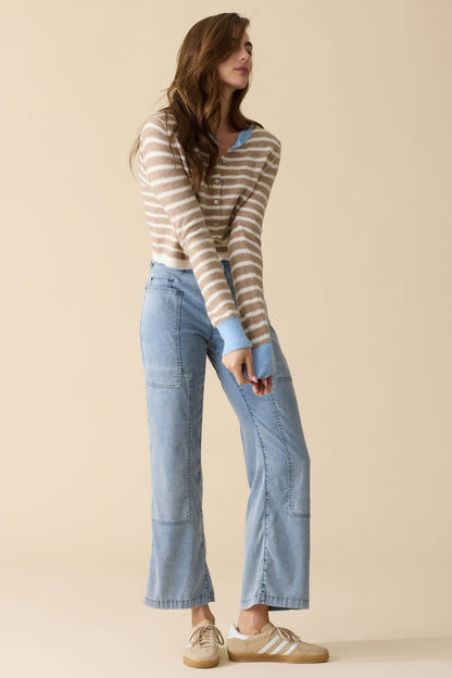 A woman modeling a beige and brown horizontally striped knit sweater with medium wash bluffton patchwork pants and beige sneakers, standing against a solid beige studio background.