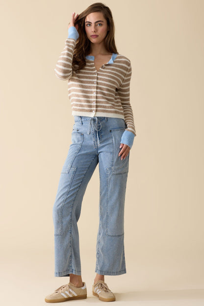 A woman modeling a beige and white striped knit cardigan with light blue trim, paired with medium wash bluffton patchwork pants and beige sneakers, against a solid beige studio background.