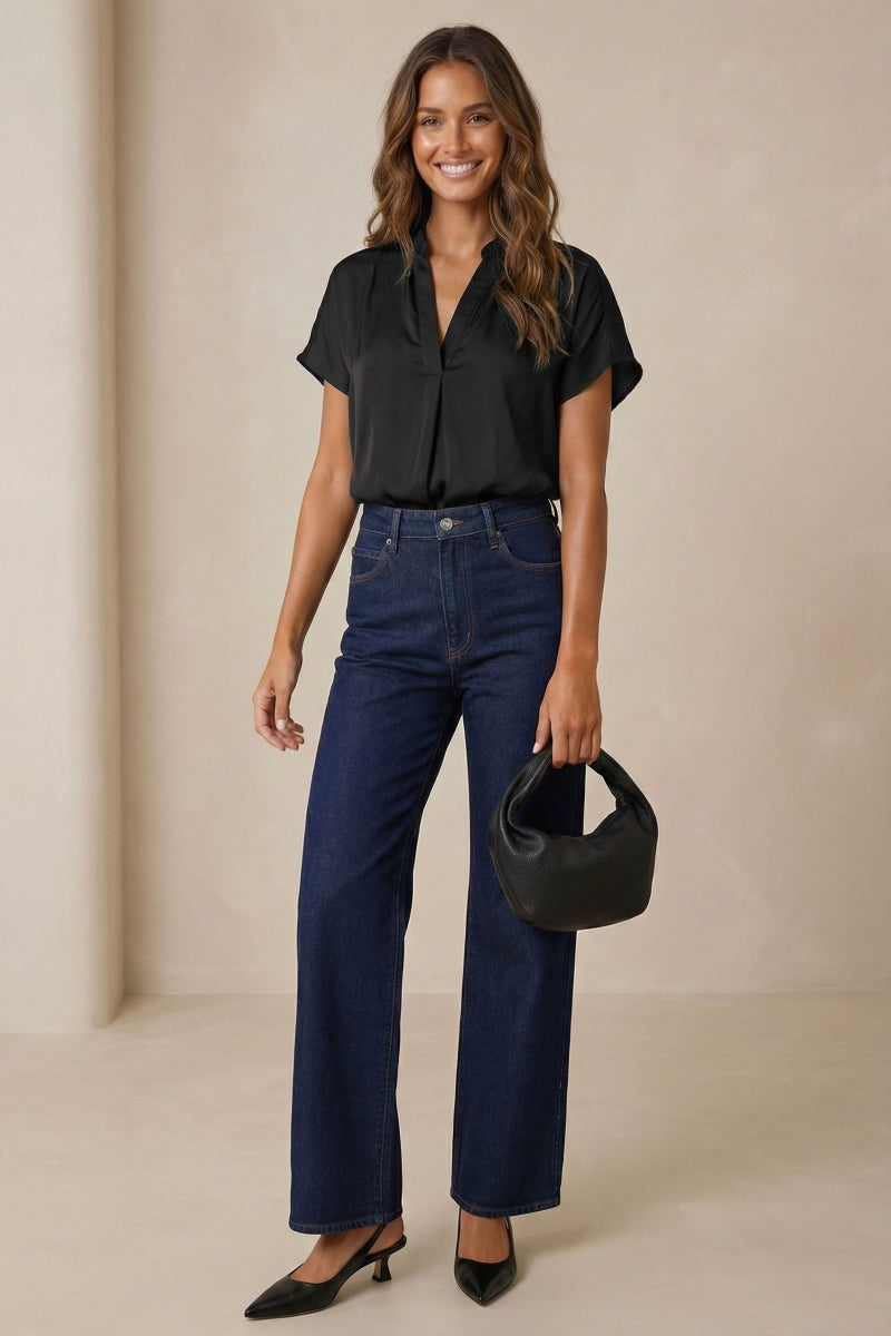 Woman wearing a drop shoulder black blouse and blue jeans holding a black handbag against a beige background